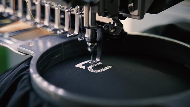 Stickmaschine stickt ein Logo auf einen Textilstoff in hoher Qualität. / Industrielle Fertigung in der Textil Fabrikation mit der Nähmaschine.