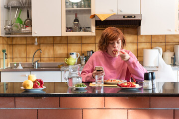 Young cute woman eats in her kitchen and uses smartphone.
