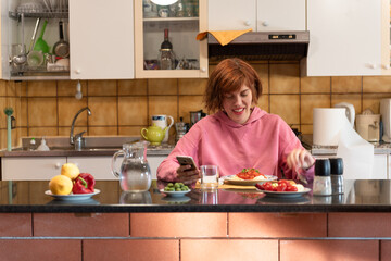 Young cute woman eats in her kitchen and uses smartphone.