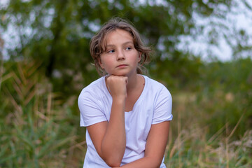 Naklejka na ściany i meble Pensive sad teenager girl looks to the side. Portrait in the park outdoors. Emotions, feelings concept. Blurred background