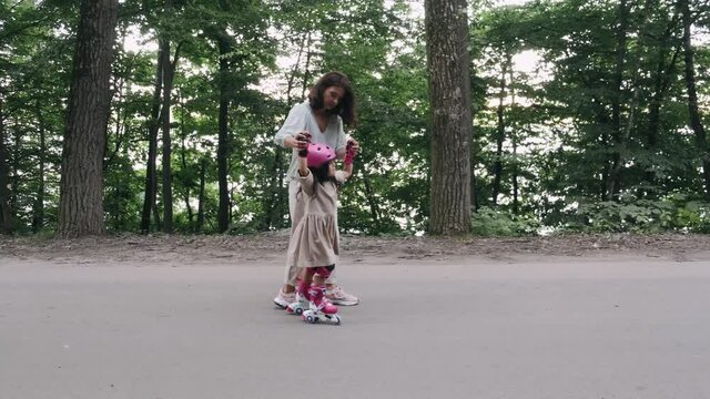 A young mother is having quality tie with daughter. The kid is rollerblading in skates and a helmet. They are in a green park.
