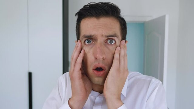 Scared businessman looks into camera with startle at face clutches his head showing fear and surprise. Portrait of shocked man on office background.