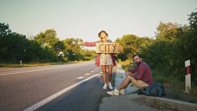 Couple of travellers hitchhiking on empty road, man resting on ground