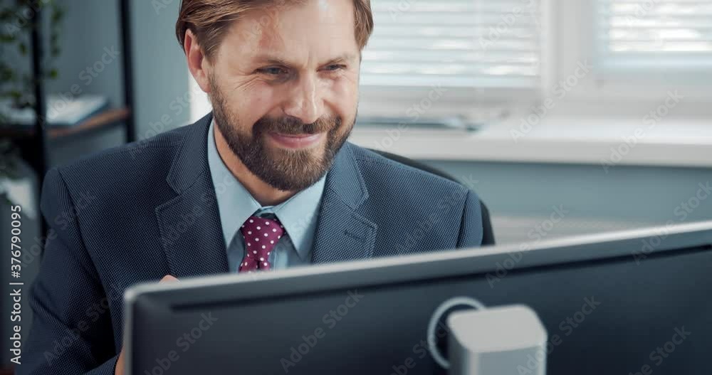 Poster handsome bearded man in formal wear smiling and gesturing while sitting at workplace with modern com