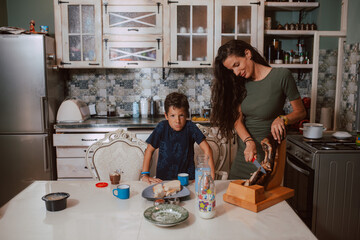 A beautiful caucasian mother in a green dress prepares breakfast for her two sons who sit at the table and have breakfast in the kitchen. Family time