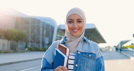 Portrait of young Arabic beautiful woman in headscarf smiling and holding textbooks and books. Pretty Arabian female student in hijab at street. Islamic happy girl. Education for Arabic girls.