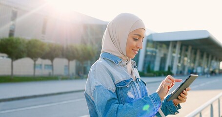 Young muslim pretty joyful woman in traditional hijab using tablet device at street. Urban...