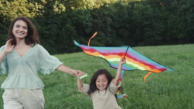 A happy parent and kid are haing fun outside on sumer holidays. The mother ad daughter are walking with a kite on the grass in a park.