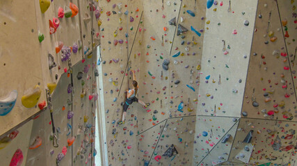 AERIAL: Climber girl climbs up an indoor climbing route filled with jug holds.
