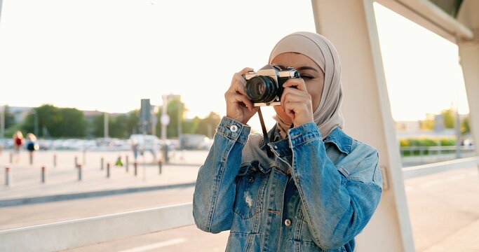 Muslim young beautiful stylish woman in traditional headscarf taking photos on vintage photo camera at street. Female in Arabic hijab making pictures while standing outdoor in city.