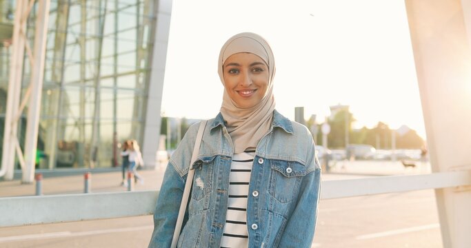 Portrait shot of beautiful happy young Arabian woman in traditional headscarf looking and smiling at camera in sunlight. Sunny summer day outdoor. Muslim cheerful female in hijab. Islamic girl in city