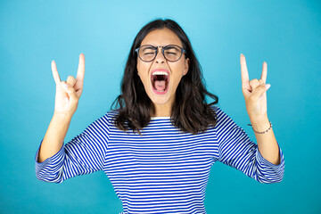 Pretty woman wearing glasses standing over insolated blue background shouting with crazy expression doing rock symbol with hands up