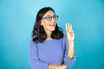 Pretty woman wearing glasses standing over insolated blue background showing and pointing up with fingers number three while smiling confident and happy.