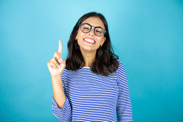 Pretty woman wearing glasses standing over insolated blue background showing and pointing up with fingers number one while smiling confident and happy.