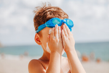 portrait of a boy in blue swimming glasses smearing his face with sun lotion
