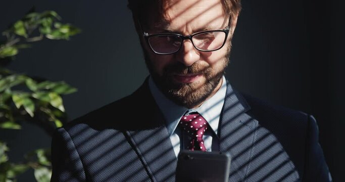 Portrait of bearded businessman in eyeglasses using modern smartphone while standing at office with shadow from jalousie on his face. Concept of people and technology.