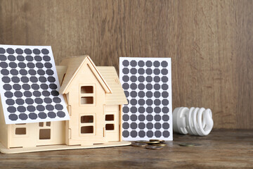 Wooden house model with solar panels, lightbulb and coins on table