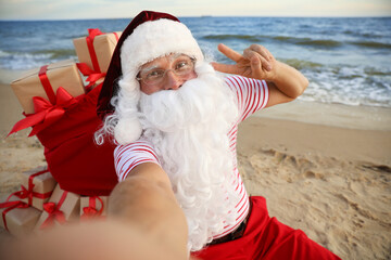 Obraz premium Santa Claus with bag of presents taking selfie on beach. Christmas vacation