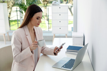 Young woman drinking coffee while using phone in office
