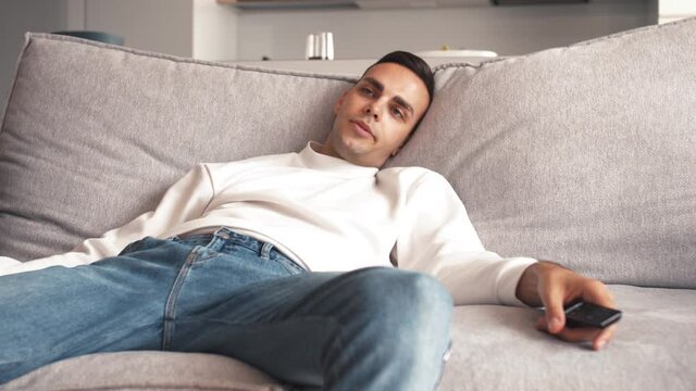 An extremely bored and frustrated caucasian man sitting on a sofa at home struggle to watch movie, changing channels with remote