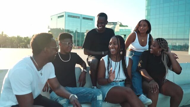 Group of young modern african black friends happily sitting together on the bench of the stairs in bright sunlight enjoying summer vacation