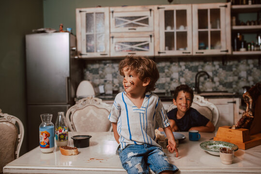 Beautiful little caucasian dirty boy sitting on the table in the kitchen, while his older brather standing behind him. Kids in the kitcen