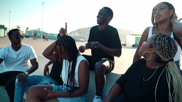 Group of young modern african black friends happily sitting together on the bench of the stairs in bright sunlight enjoying summer vacation