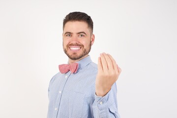 European man dressed in formal shirt and bow tie poses against white studio background inviting to come with hand. Happy that you came