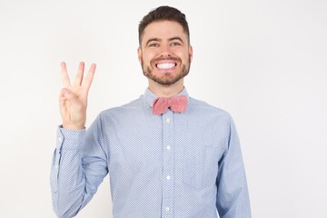 European man dressed in formal shirt and bow tie poses against white studio background showing and pointing up with fingers number three while smiling confident and happy.
