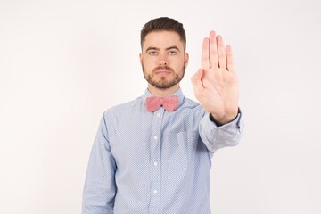 Fototapeta premium European man dressed in formal shirt and bow tie poses against white studio background doing stop gesture with palm of the hand. Warning expression with negative and serious gesture on the face