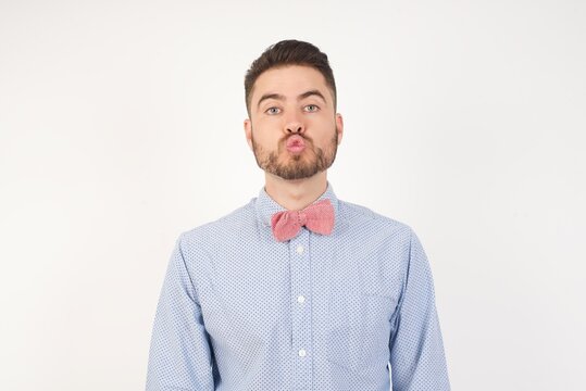 Shot of pleasant looking European man dressed in formal shirt and bow tie poses against white background, pouts lips, looks with green eyes at camera,  has fun with girlfriend,