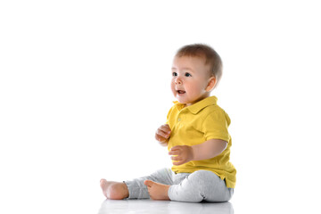 small baby in a yellow polo sits on the floor, laughs, touches his legs and looks to the side at the top corner on a white background.