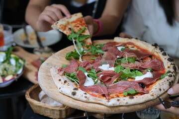close up hand taking pizza with cured meats, arugula and cheese. On wooden plate. Defocused background