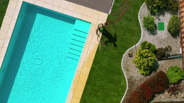 Aerial Image Of Male Landscaping Worker Watering Narrow Strip Of Newly Rolled Sod In Secluded Backyard Of House. 