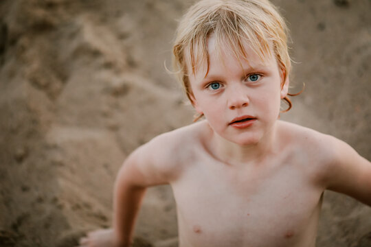 Little boy sitting in sand gazing at camera. Shirtless and wet after swimming at the beach