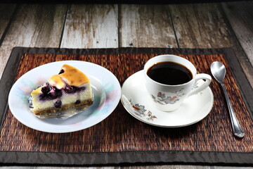 Traditional supper menu set on the wooden mat, piece of blueberry cheesecake on the plate serving with hot black coffee in the cup. Famous sweet dessert menu in bakery or cafe shop. 