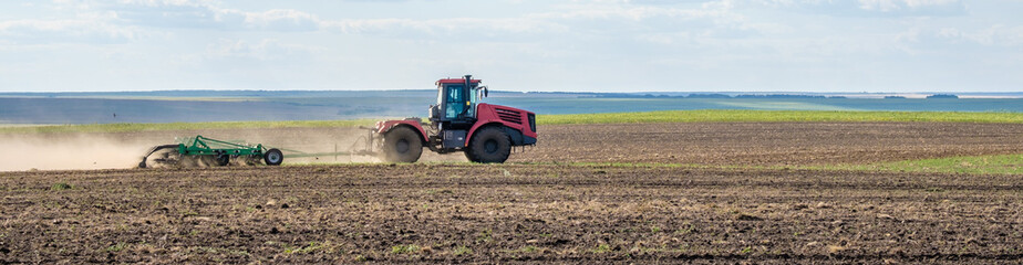 A red farm tractor in a cloud of dust cultivates the soil in the field with a cultivator after harvest. Summer sunny day. Fertile land. Modern agricultural machinery. Copy space. Banner.