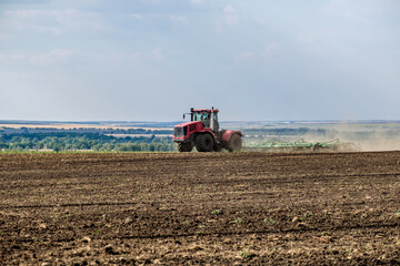 A red farm tractor in a cloud of dust cultivates the soil in the field with a cultivator after...