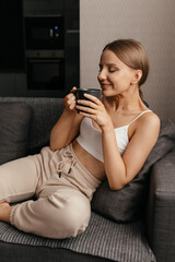 beautiful woman in top and pants sits on the sofa with a mug of tea and sniffs its delicious aroma. Positive emotions.