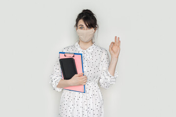 Charming brunette woman wearing a medical mask and summer dress is gesturing hello on a white studio wall holding some folders
