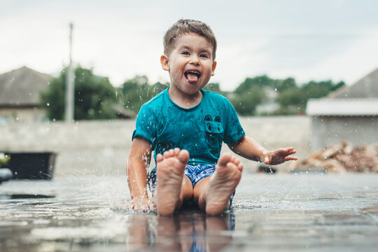 Front view photo of a caucasian boy gesturing happiness with tongue while playing barefoot in water on the ground after the rain