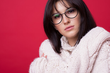 Close up photo of a caucasian brunette woman with eyeglasses posing in a warm sweater on a red studio wall embracing herself