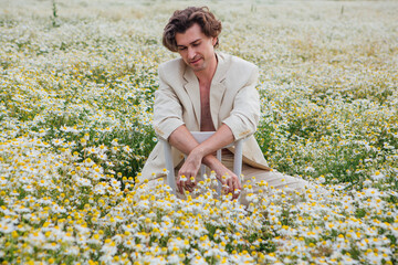 Naklejka na ściany i meble Tall handsome man sitting on a white chair in camomile flowers field