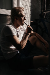 young man smokes an electronic cigarette at home on the balcony in harsh sunlight. Smoke and steam...