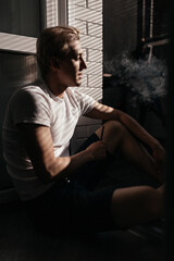 young man smokes an electronic cigarette at home on the balcony in harsh sunlight. Smoke and steam...
