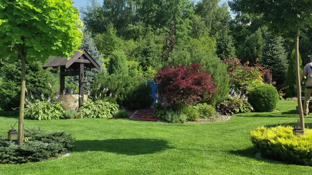 Backyard Landscape Design With Multicolored Shrubs Plants And Well Surrounded By Bright Green Lawn. Male Worker Mowing Grass. 