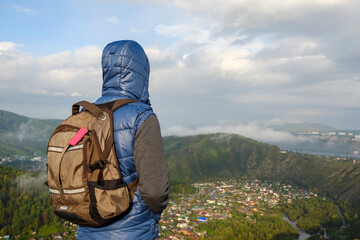 A man at the top of the mountain. Hiking backpack, hood. View from the back. Background - a valley in the mountains, residential buildings, a river, fog. Sky with clouds. Tourism, hiking concept.