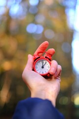 Autumn season .Autumn time. Red alarm clock in hand on blurred autumn  forest background.Symbol of...