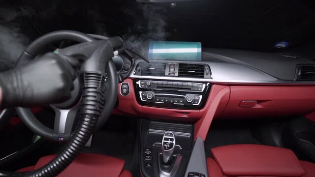 Mechanic wearing a black rubber glove and holding a hot steam vacuum cleaner cleaning the vents of the air conditioning system in a luxurious red leather car interior. Cleaning luxury car interior .