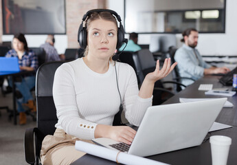 Focused young female freelancer wearing headset discussing new project with her partner online in open plan office